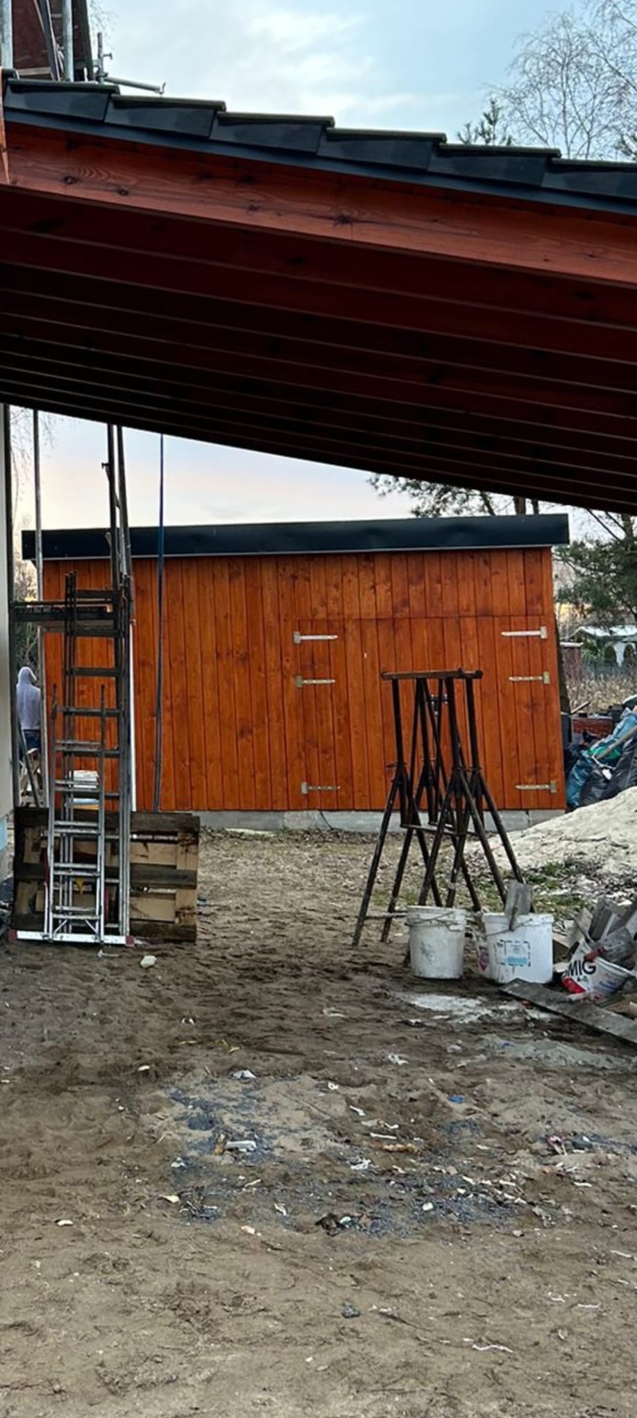 Nowo postawiony, drewniany budynek gospodarczy z dwuskrzydłowymi drzwiami, widoczny z boku, z elementami rusztowania i narzędzi budowlanych na pierwszym planie.