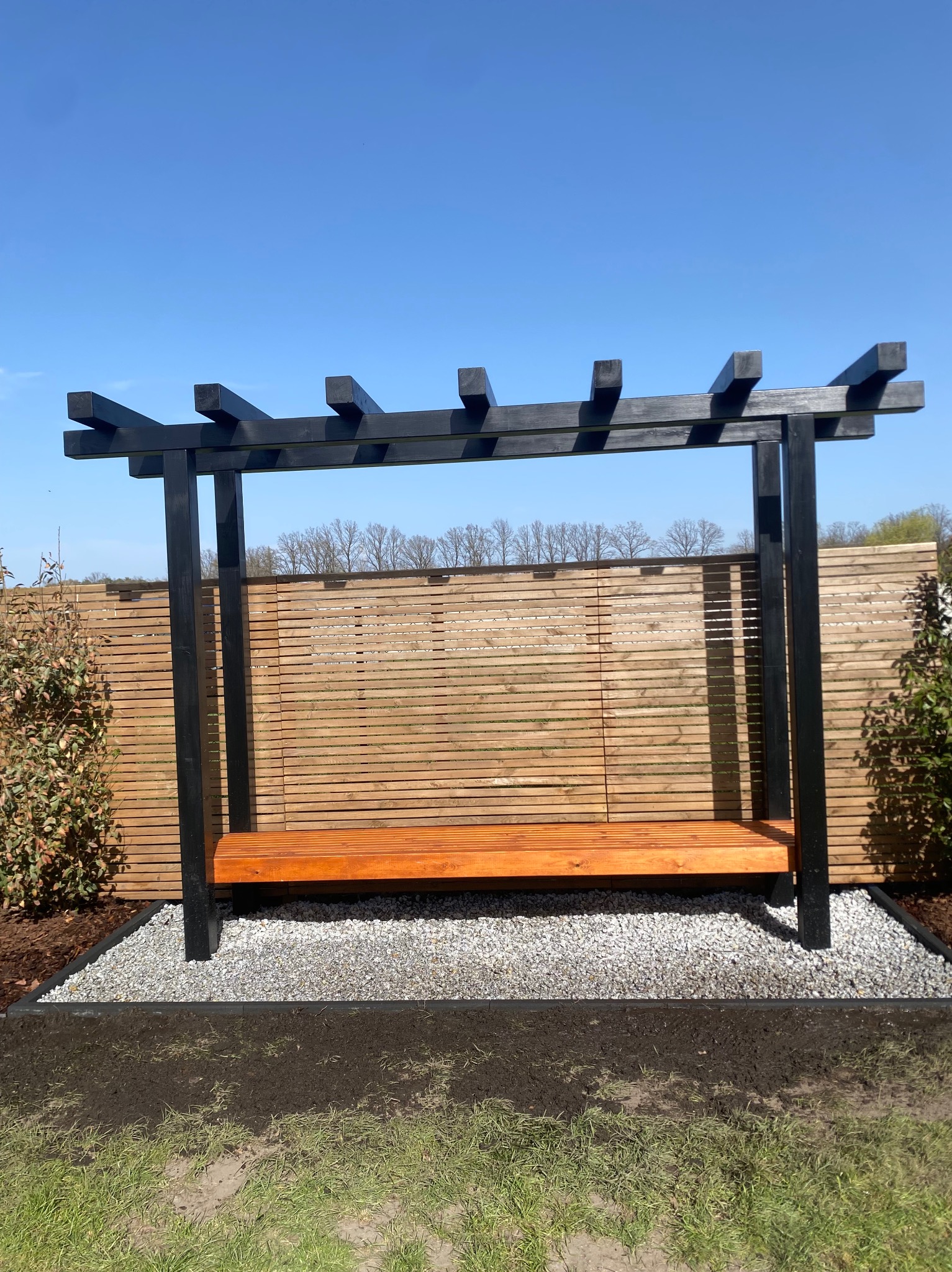 Nowoczesna pergola z drewnianą ławką, pomalowana na czarno, na tle drewnianego ogrodzenia, otoczona białym żwirem i świeżo posadzoną zielenią.
