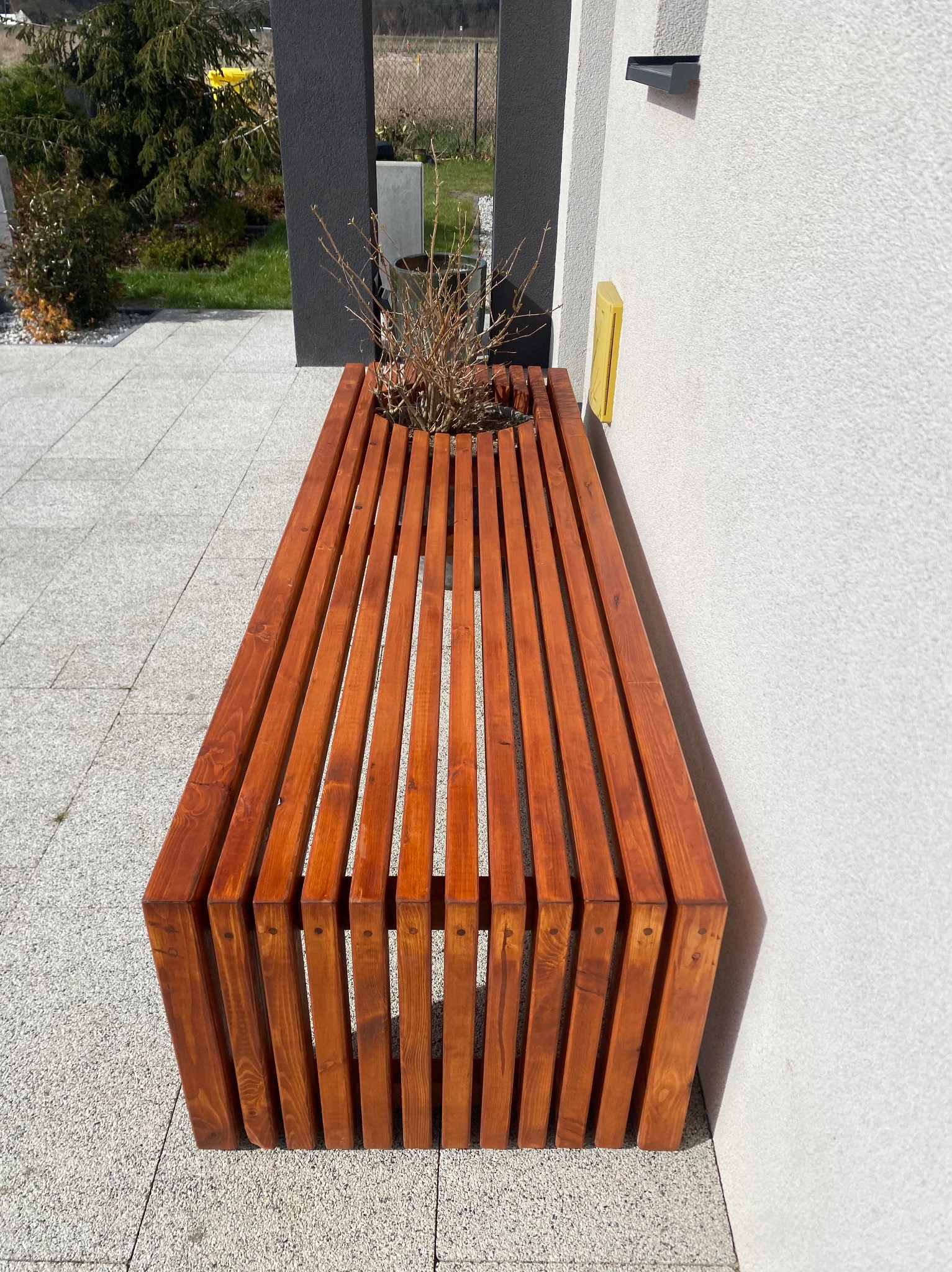 Drewniana ławka z wbudowaną donicą, wykonana z pionowych listew w ciepłym odcieniu brązu, umieszczona na szarym bruku w pobliżu białej ściany budynku.