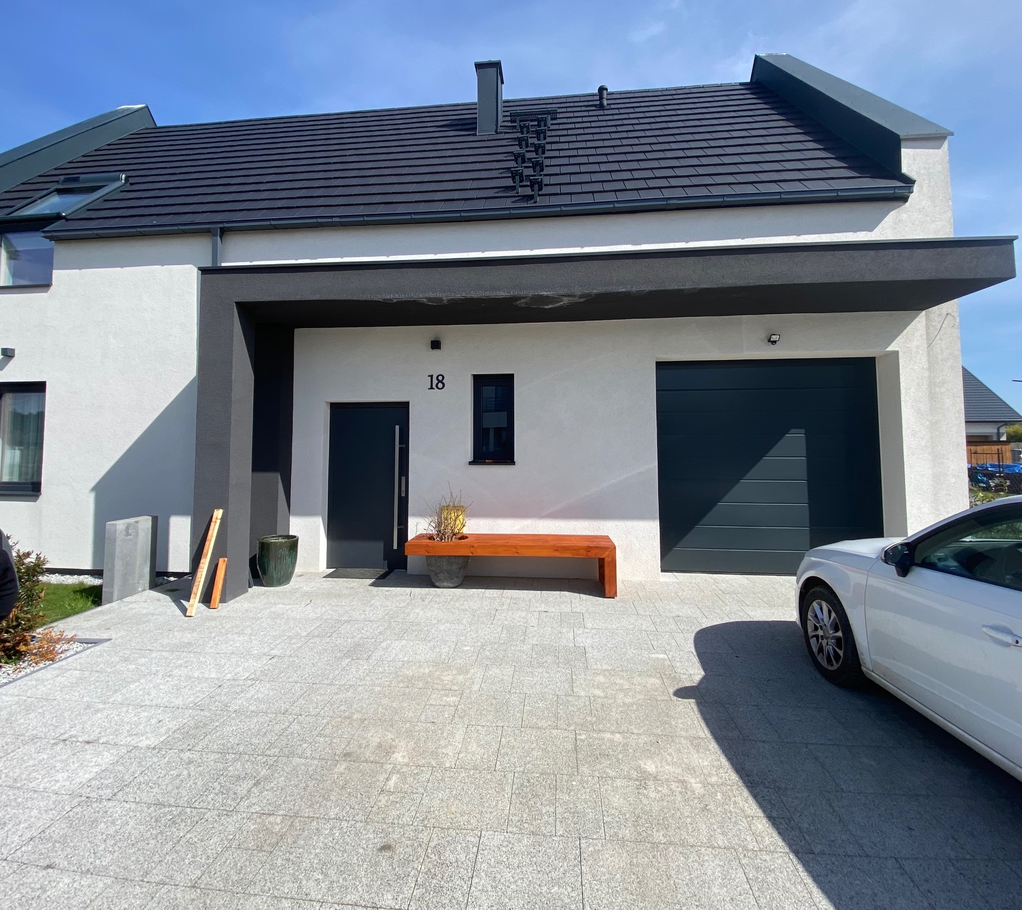 Nowoczesny dom jednorodzinny z szarą elewacją, ciemną stolarką okienną i garażem, drewnianą ławką z donicą przed wejściem, oraz podjazdem wyłożonym jasnoszarą kostką brukową.