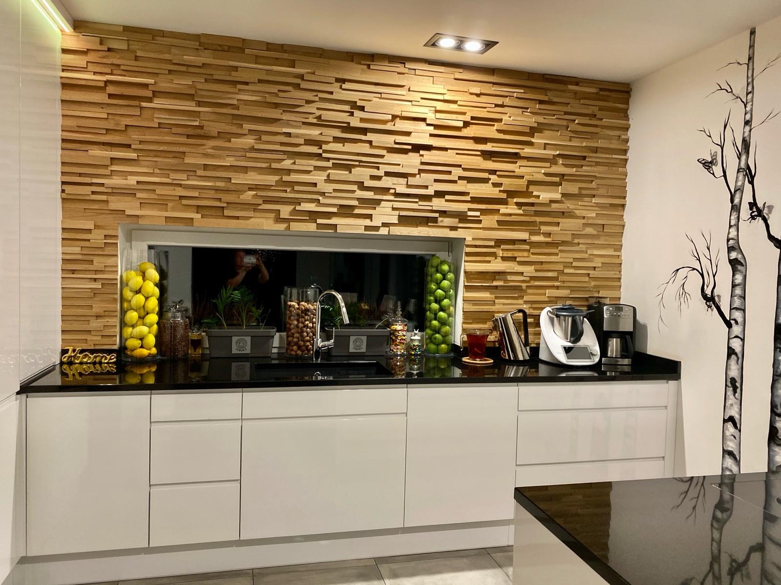 Nowoczesna kuchnia z dekoracyjną ścianą z drewnianych paneli, czarnym blatem, białymi szafkami, szklanymi pojemnikami z cytrynami i jabłkami oraz motywem brzozy na ścianie.
