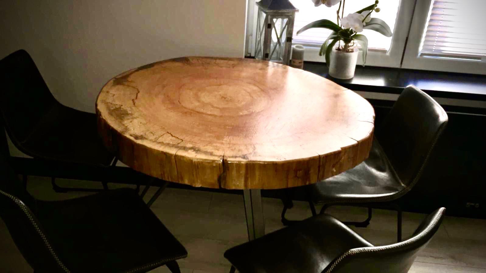 Okrągły stół z blatem z plastra drewna na metalowej nodze, otoczony czterema czarnymi krzesłami w pomieszczeniu z oknem i dekoracjami.