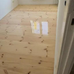 Jasna, świeżo polakierowana podłoga z desek sosnowych w pokoju, z widocznym grzejnikiem na ścianie i odbiciem okna w lakierowanej powierzchni.