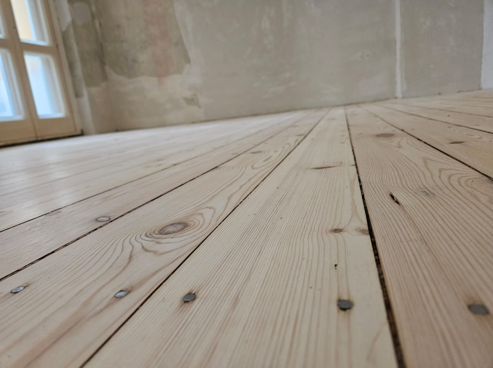 Jasne, świeżo położone deski podłogowe z widocznymi gwoździami, niedokończone ściany w tle, widok z niskiej perspektywy.