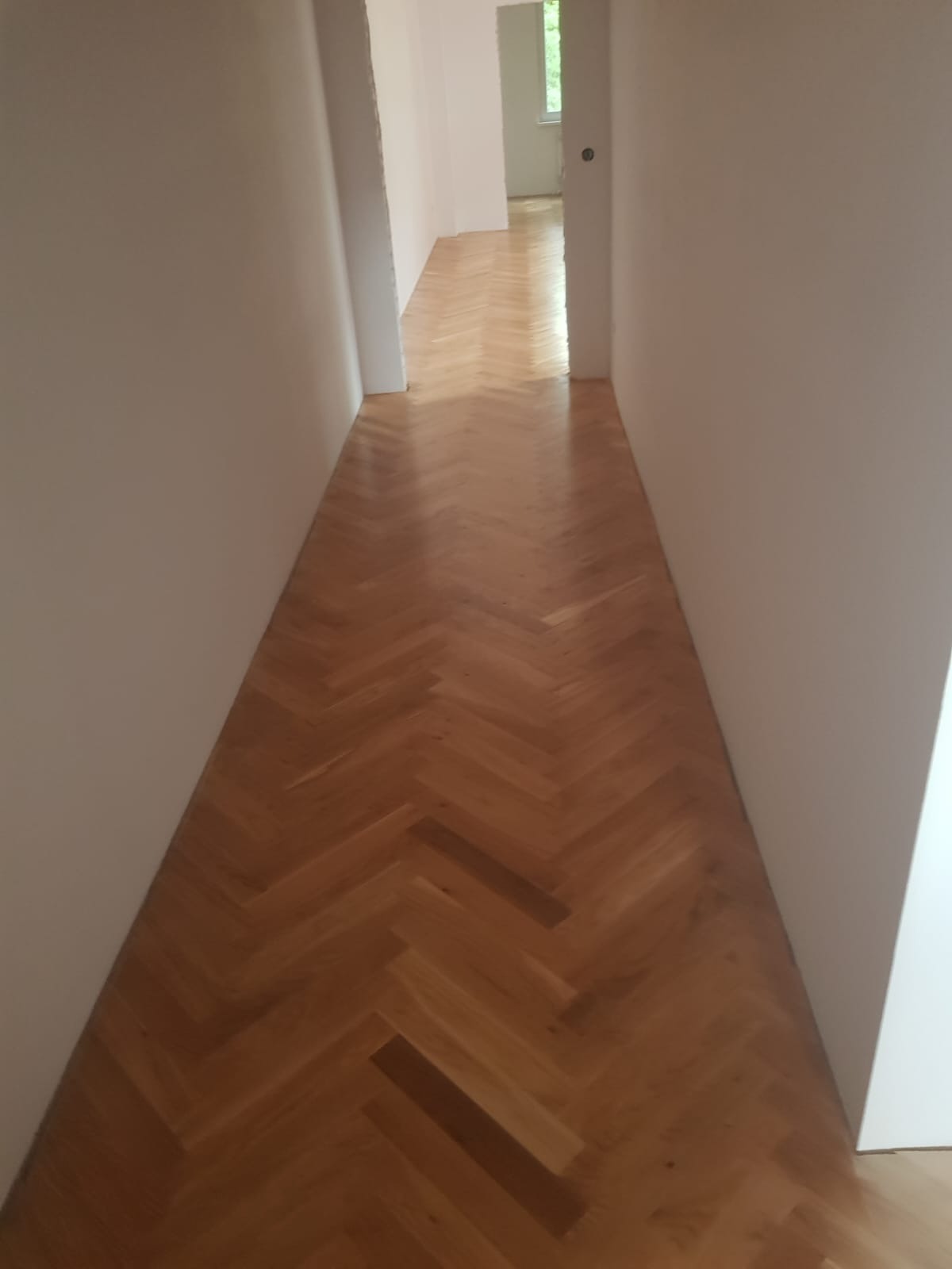 Korytarz z nową, jasną parkietową podłogą ułożoną we wzór jodełki klasycznej, widok w głąb pomieszczeń z otwartymi drzwiami.