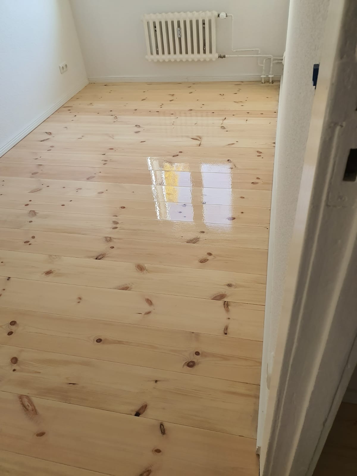 Jasna, świeżo polakierowana podłoga z desek sosnowych w pokoju, z widocznym grzejnikiem na ścianie i odbiciem okna w lakierowanej powierzchni.