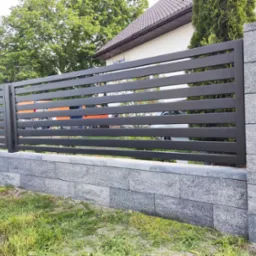 Nowoczesne, poziome przęsła z ciemnoszarego metalu tworzą ogrodzenie na podmurówce z szarych, betonowych bloczków. W tle widoczny dom z brązowym dachem i zielone drzewa.