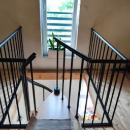 Metalowa balustrada schodowa w kolorze czarnym, widok z góry na drewniane schody i okno z rośliną. Minimalistyczny design i jasne wnętrze.