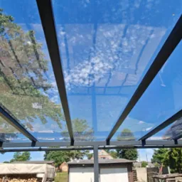 Szklana pergola z czarną metalową konstrukcją, widok z dołu na błękitne niebo z chmurami i zieleń drzew. Nowoczesny design i funkcjonalność zadaszenia.