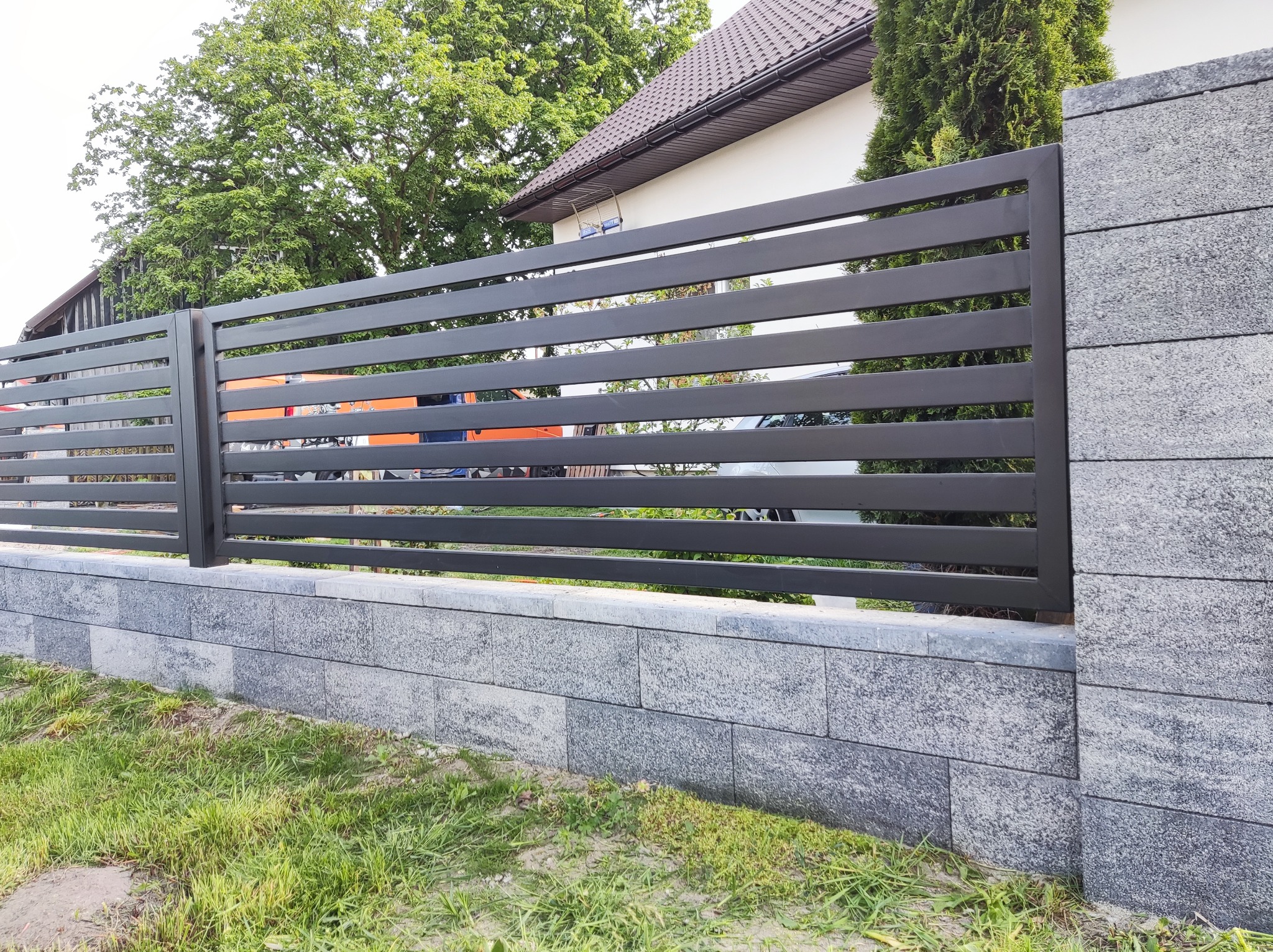 Nowoczesne, poziome przęsła z ciemnoszarego metalu tworzą ogrodzenie na podmurówce z szarych, betonowych bloczków. W tle widoczny dom z brązowym dachem i zielone drzewa.