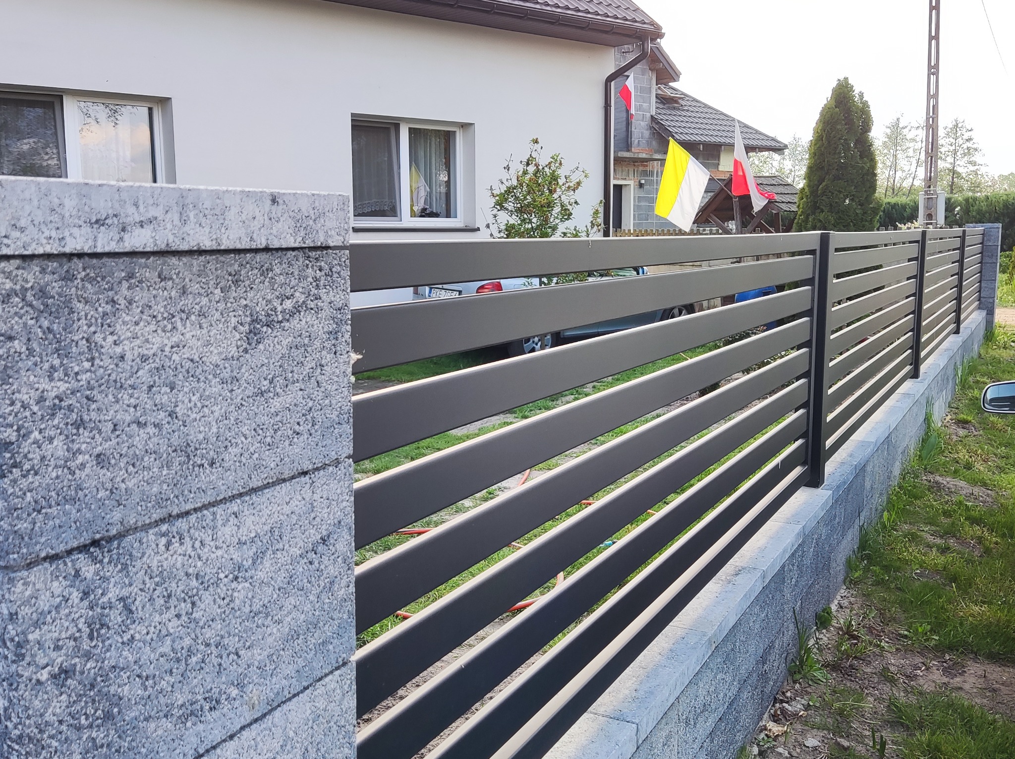 Nowoczesne ogrodzenie panelowe z poziomych, szarych profili zamontowane na niskiej podmurówce z szarego kamienia łupanego, widok na dom z polską i watykańską flagą w oknach.