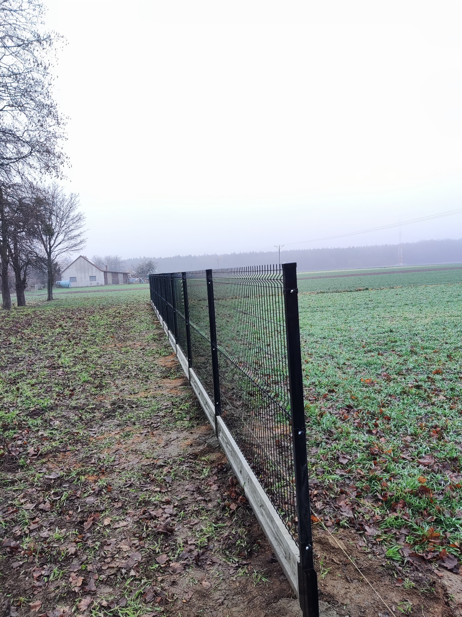 Długi, czarny panelowy płot z podmurówką ciągnący się wzdłuż pola w pochmurny dzień. W tle widoczny budynek gospodarczy i las.