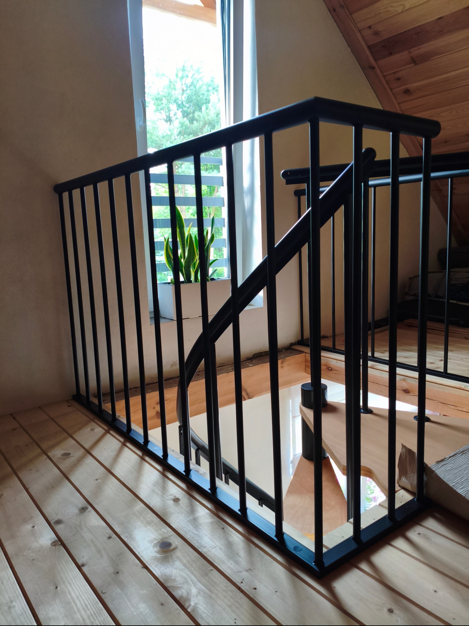Czarna, metalowa balustrada schodowa w nowoczesnym wnętrzu z drewnianą podłogą i oknem z rośliną w tle, widziana z dołu.