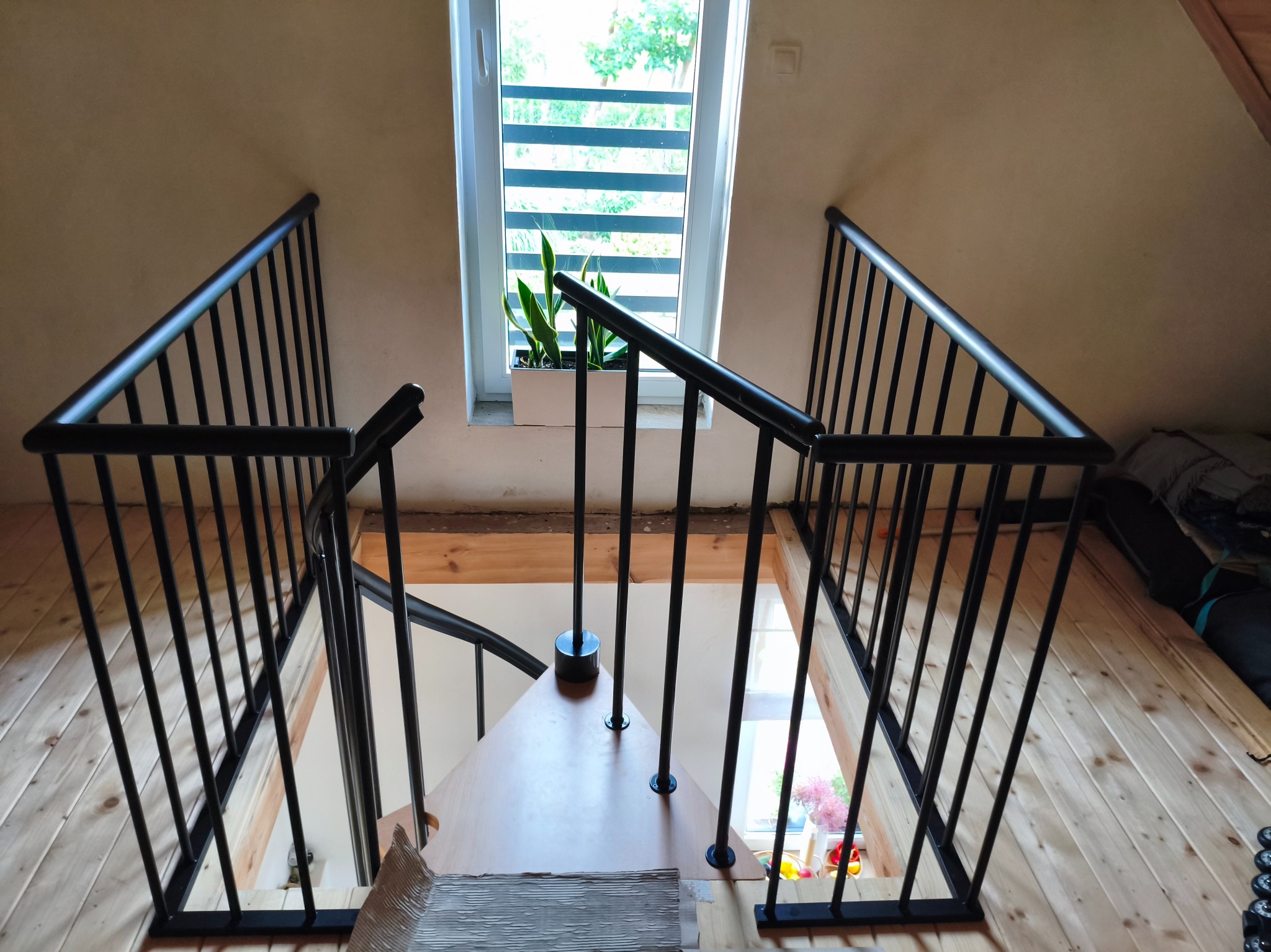 Metalowa balustrada schodowa w kolorze czarnym, widok z góry na drewniane schody i okno z rośliną. Minimalistyczny design i jasne wnętrze.