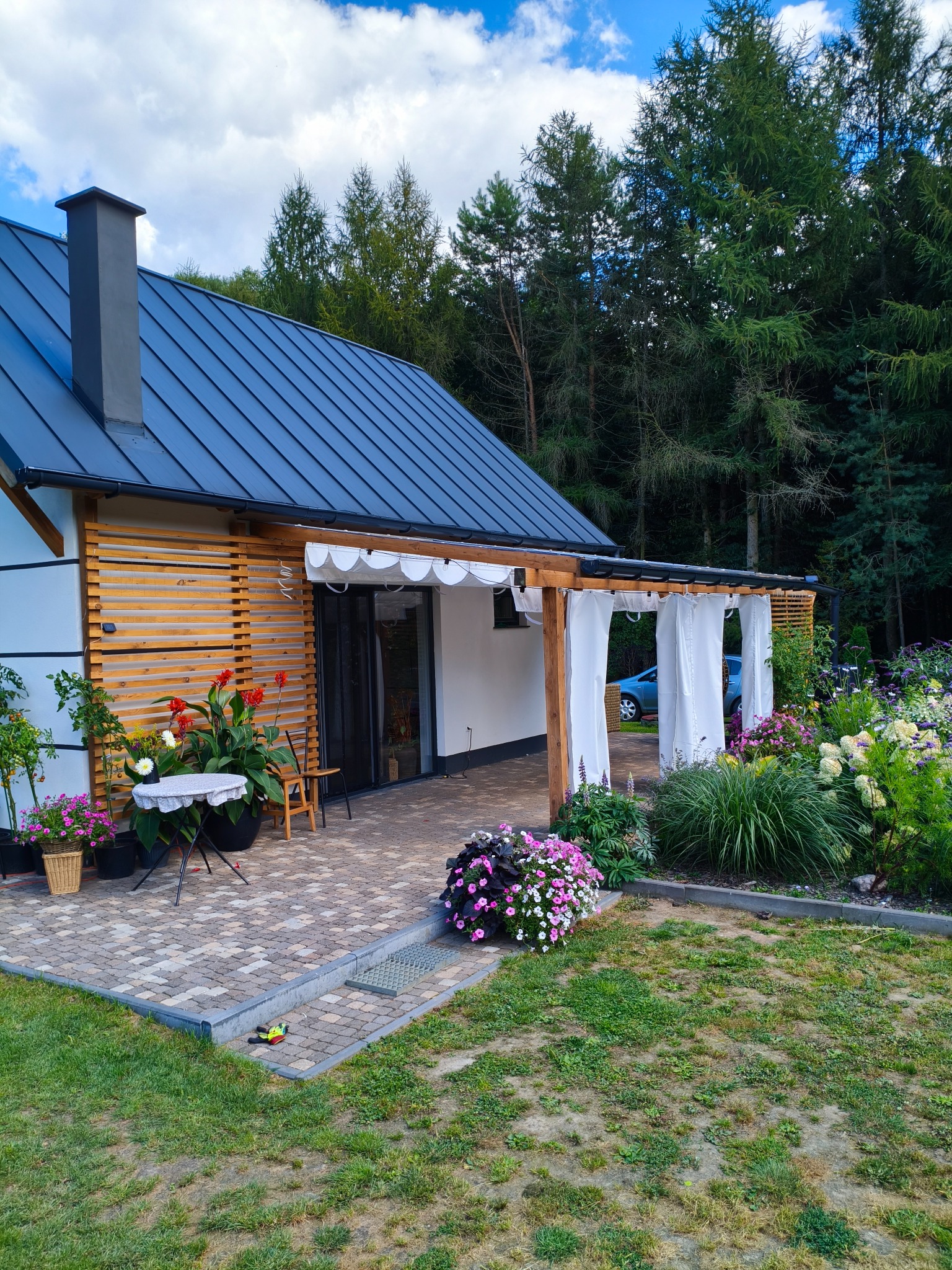 Drewniana pergola z białymi zasłonami przy nowoczesnym domu. Umeblowany taras z kwiatami, zadbany ogród, las w tle. Detal: zabawka na kostce brukowej.