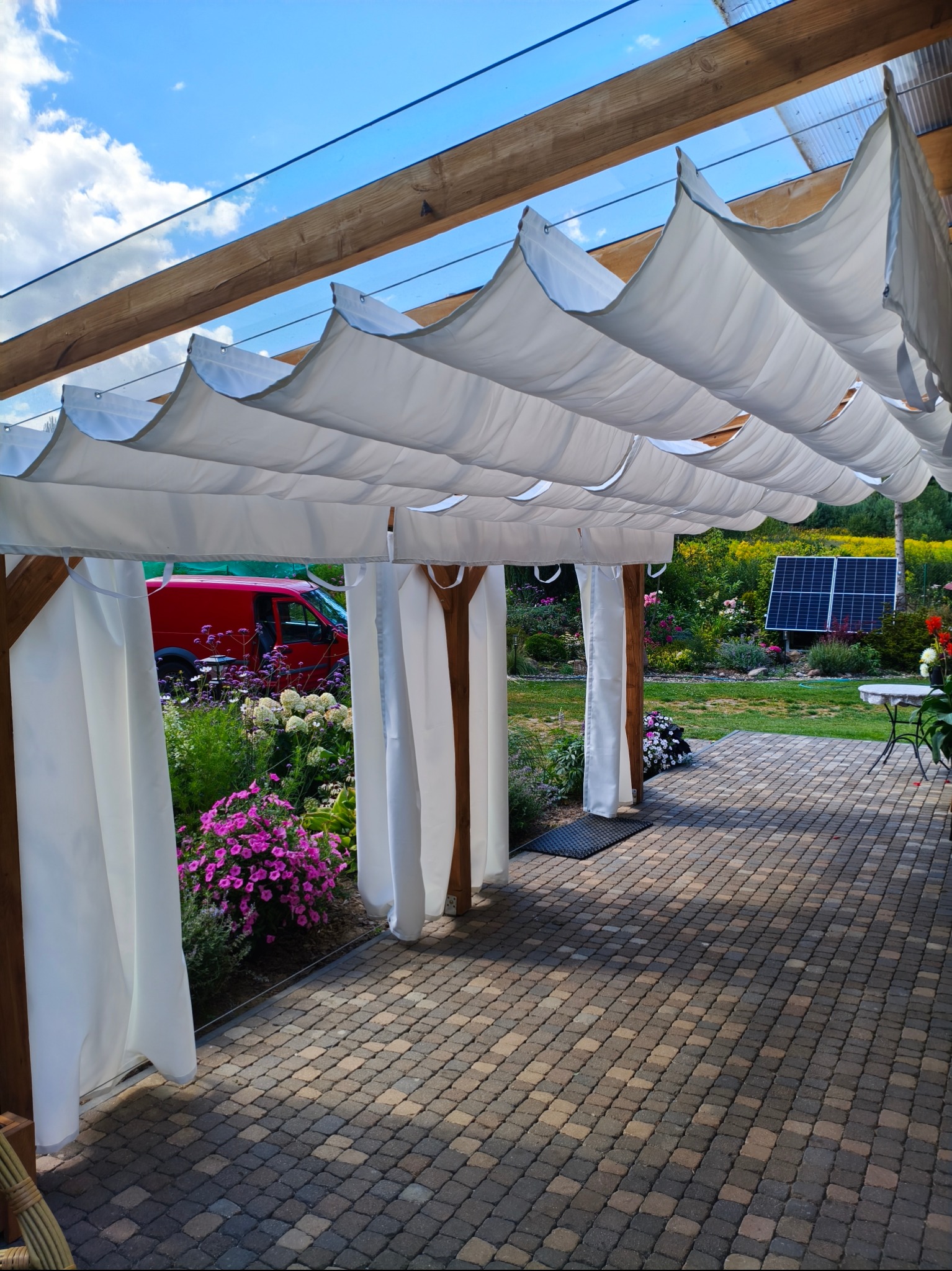 Drewniana pergola z białymi, falującymi zasłonami i przezroczystym dachem, widok na ogród z panelem słonecznym i czerwonym samochodem w tle. Brukowana posadzka.