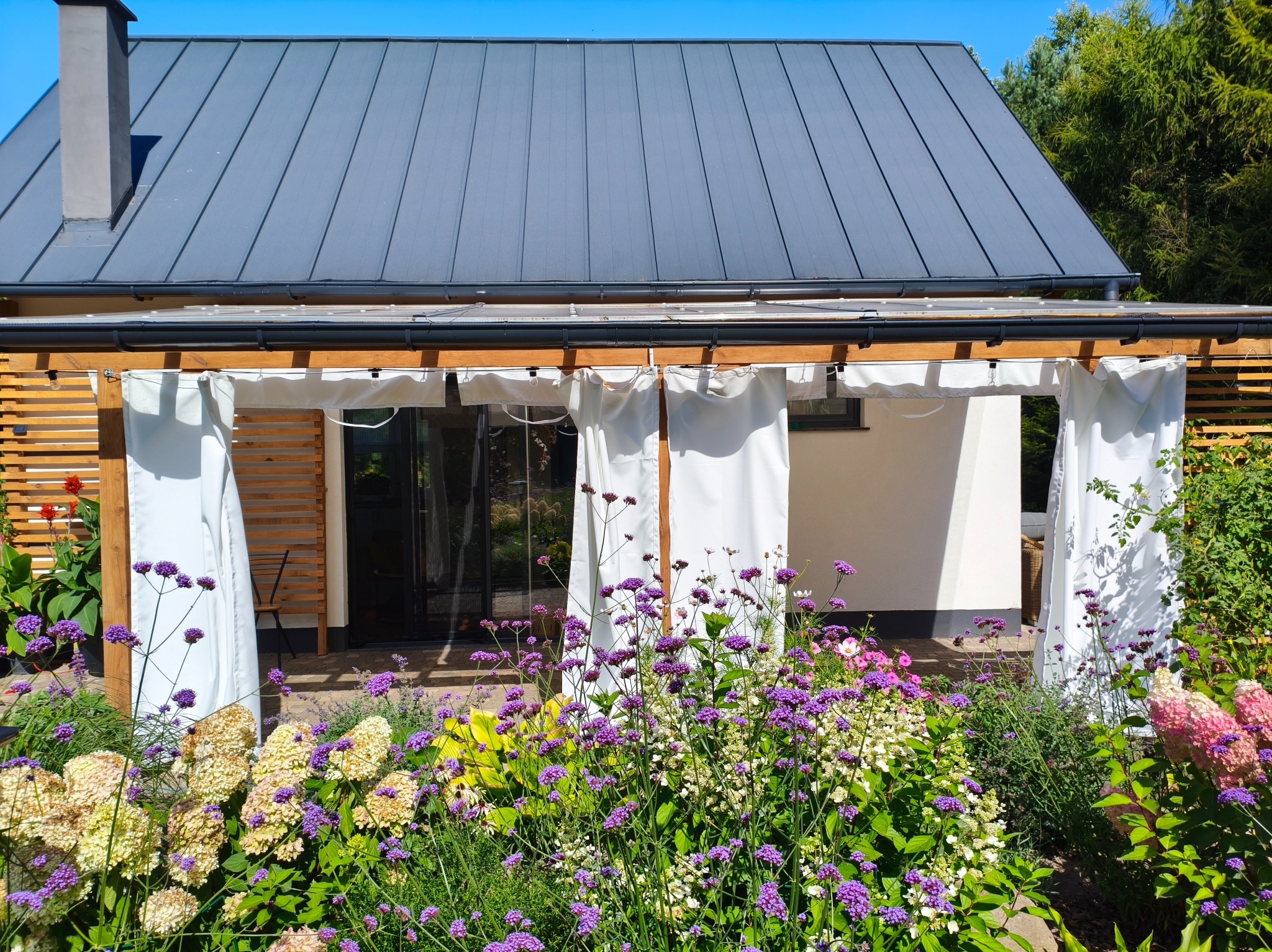 Pergola z białymi zasłonami i drewnianą konstrukcją, otoczona kolorowym ogrodem, na tle domu z szarym dachem. Urokliwe zacisze w słoneczny dzień.