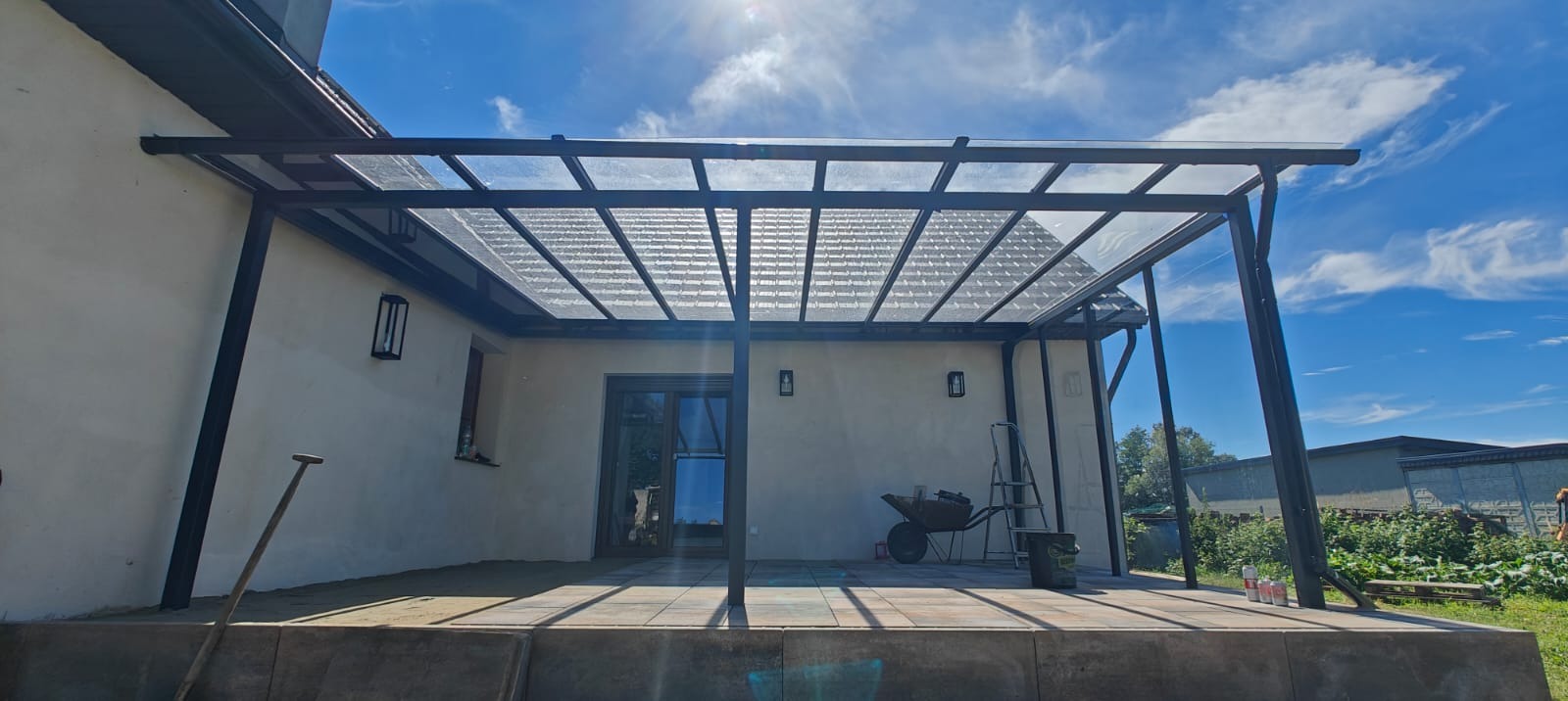 Metalowa pergola z przezroczystym zadaszeniem przylegająca do budynku, widok na ogród w słoneczny dzień. Na tarasie narzędzia: taczka, drabina i puszki z farbą.