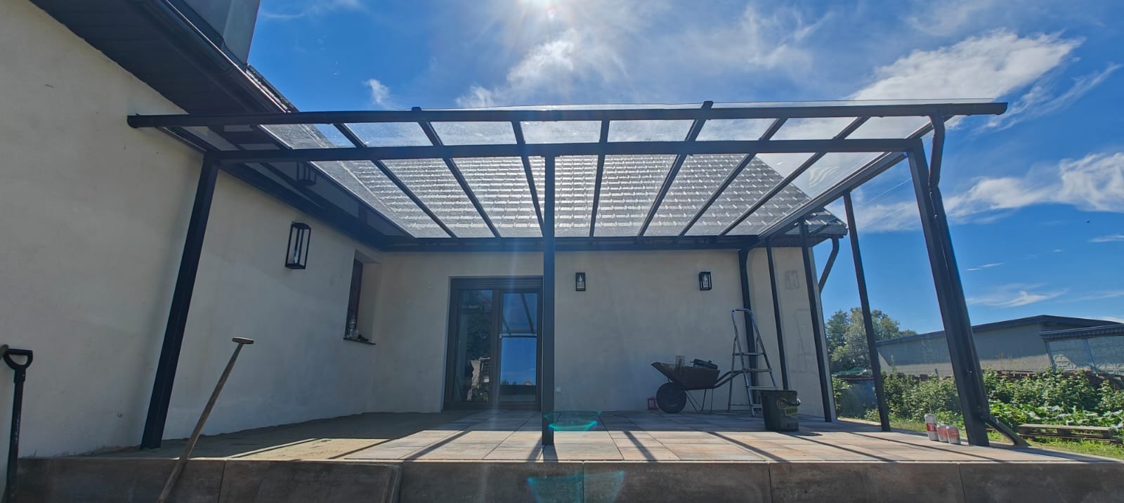 Nowoczesna pergola metalowa z przezroczystym zadaszeniem, przylegająca do jasnego budynku. Widoczne narzędzia i elementy wykończeniowe na tarasie z kostki.