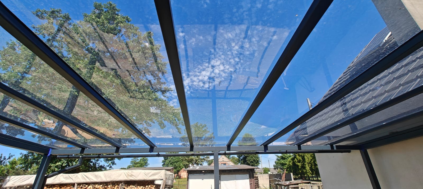 Szklana pergola z czarną metalową konstrukcją, widok z dołu na błękitne niebo z chmurami i zieleń drzew. Nowoczesny design i funkcjonalność zadaszenia.