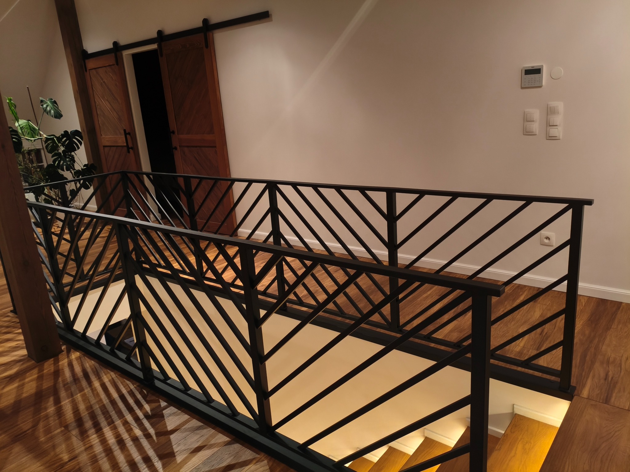 Nowoczesna, czarna balustrada schodowa z geometrycznym wzorem na drewnianych schodach i podłodze. Minimalistyczny design, ciepłe oświetlenie, widok z góry.