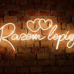 Neonowy napis 'Razem lepiej' z dwoma sercami, umieszczony na tle ściany z czerwonej cegły, oświetlony ciepłym światłem.