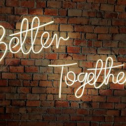 Hubert Ploch Media - Neonowy napis 'Better Together' na tle ściany z czerwonej cegły, oświetlony ciepłym światłem, tworzący romantyczną atmosferę.