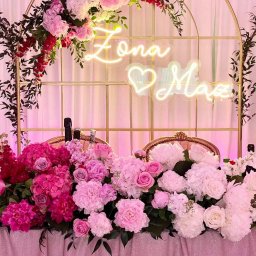 Hubert Ploch Media - Dekoracja stołu weselnego z kwiatami w odcieniach różu i bieli, neonowym napisem 'Żona ❤️ Mąż' na złotym łuku, butelki szampana w tle.