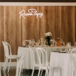 Uroczysty stół weselny z białymi krzesłami, nakryciem, kwiatami w wazonie i dekoracyjnym neonem 'Razem Lepiej' na drewnianym tle.