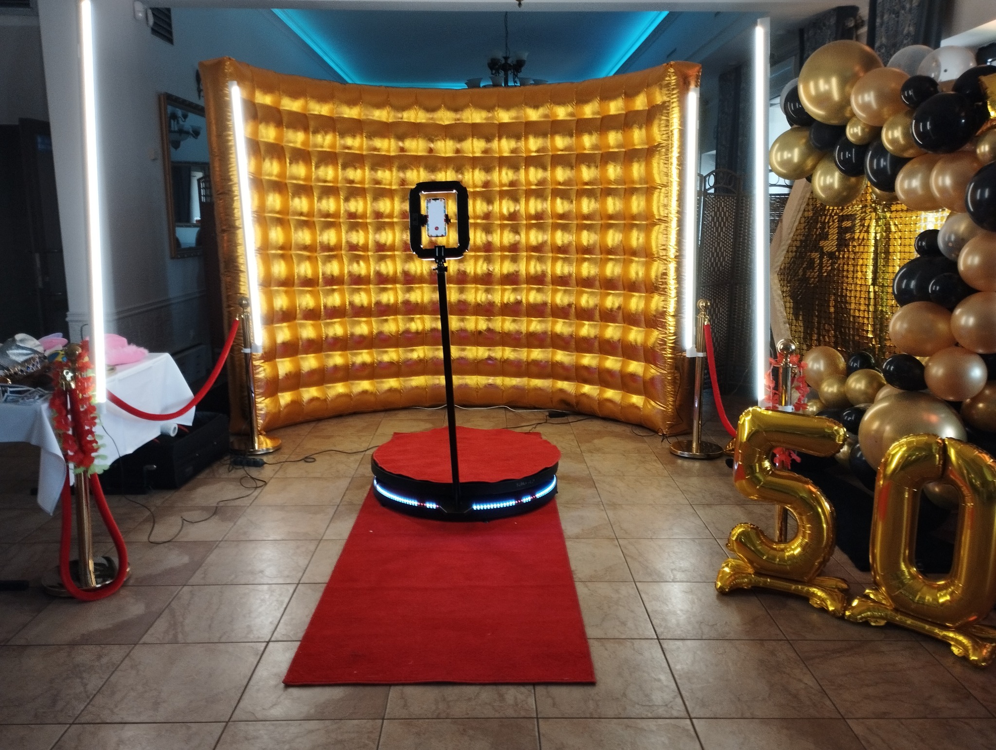 Złota, dmuchana ścianka z kwadratowymi wgłębieniami, okrągła platforma z podświetleniem LED na czerwonym dywanie, statyw z telefonem, dekoracja z balonów w kolorach złota i czerni, oraz balony...