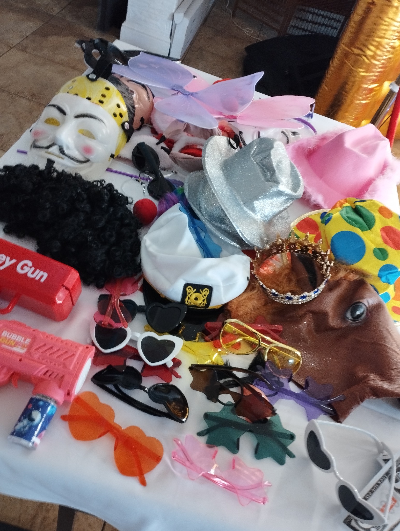 Akcesoria do przebrania na białym stole: maski, peruki, kapelusze, okulary w kształcie serc, korona, zabawkowy pistolet na bańki.