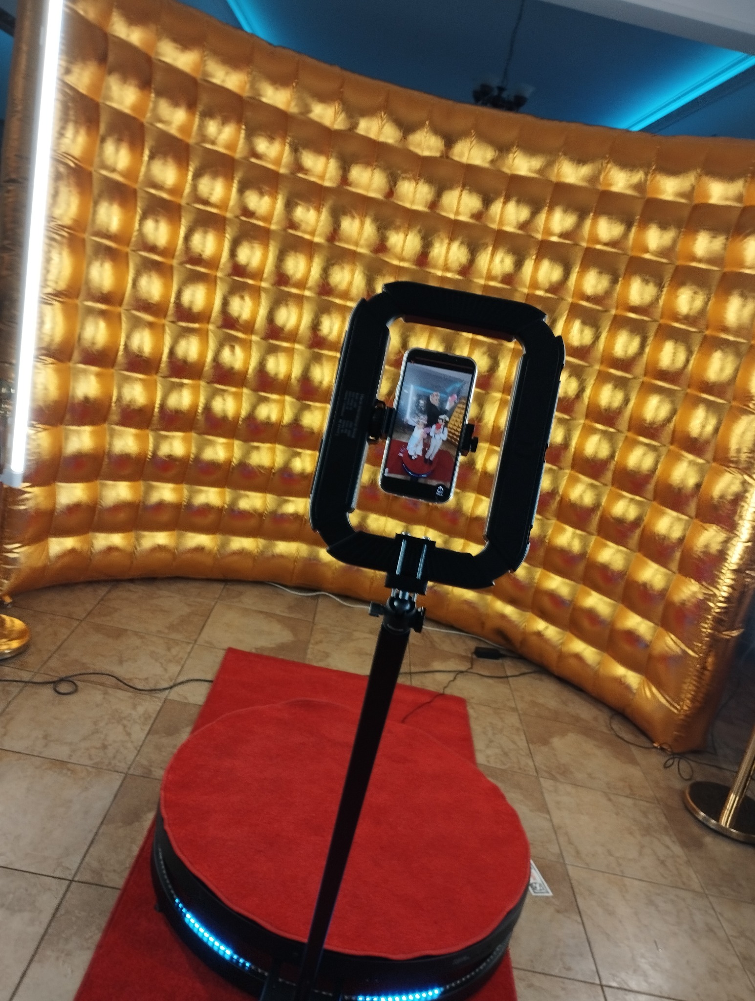 Złota, dmuchana ścianka tła, czerwony dywan i obrotowa platforma z oświetleniem LED, na której stoi statyw z telefonem, na ekranie którego widać parę młodą.