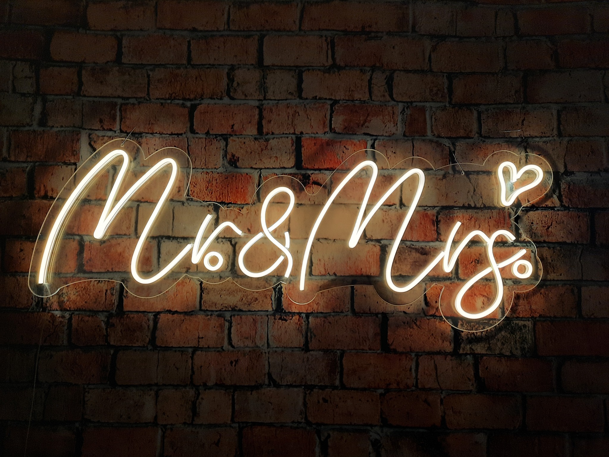 Neonowy napis 'Mrs & Mrs' z sercem, umieszczony na tle ceglanej ściany, ciepłe oświetlenie.