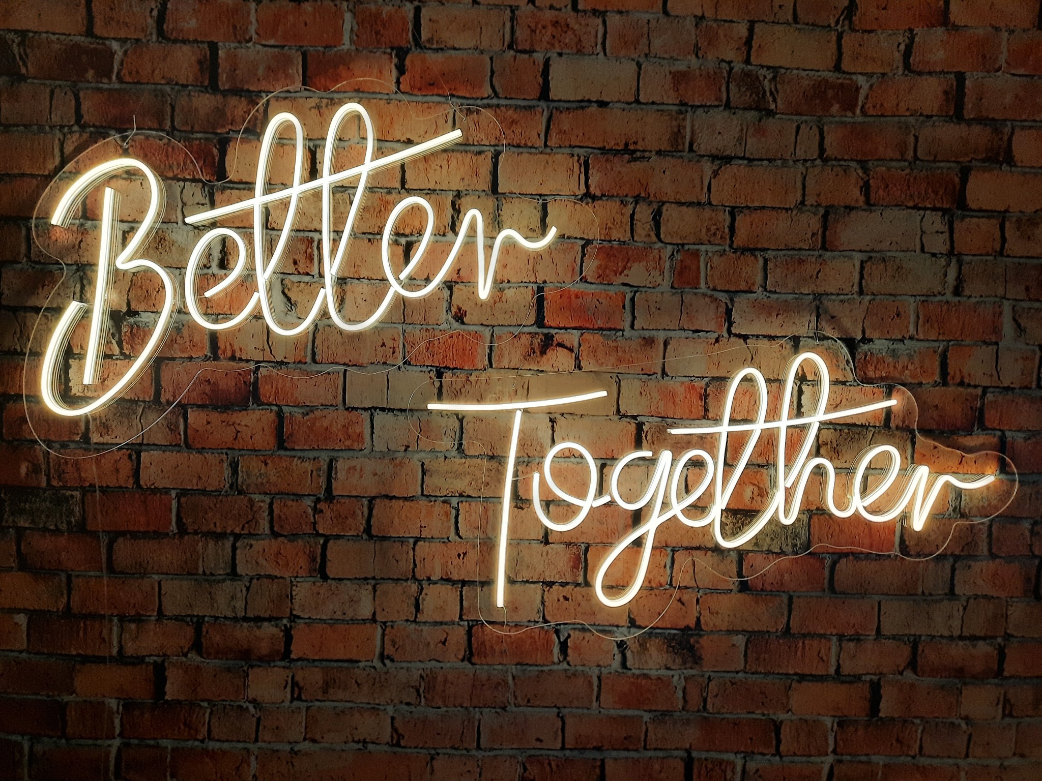 Neonowy napis 'Better Together' na tle ściany z czerwonej cegły, oświetlony ciepłym światłem, tworzący romantyczną atmosferę.