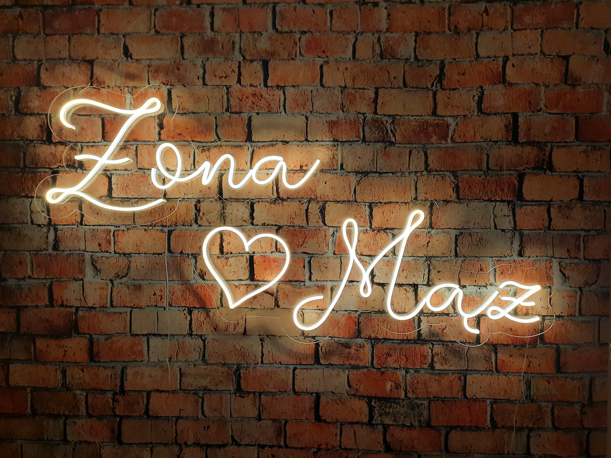 Neonowy napis 'Żona Mąż' z sercem pośrodku, umieszczony na tle ściany z czerwonej cegły.