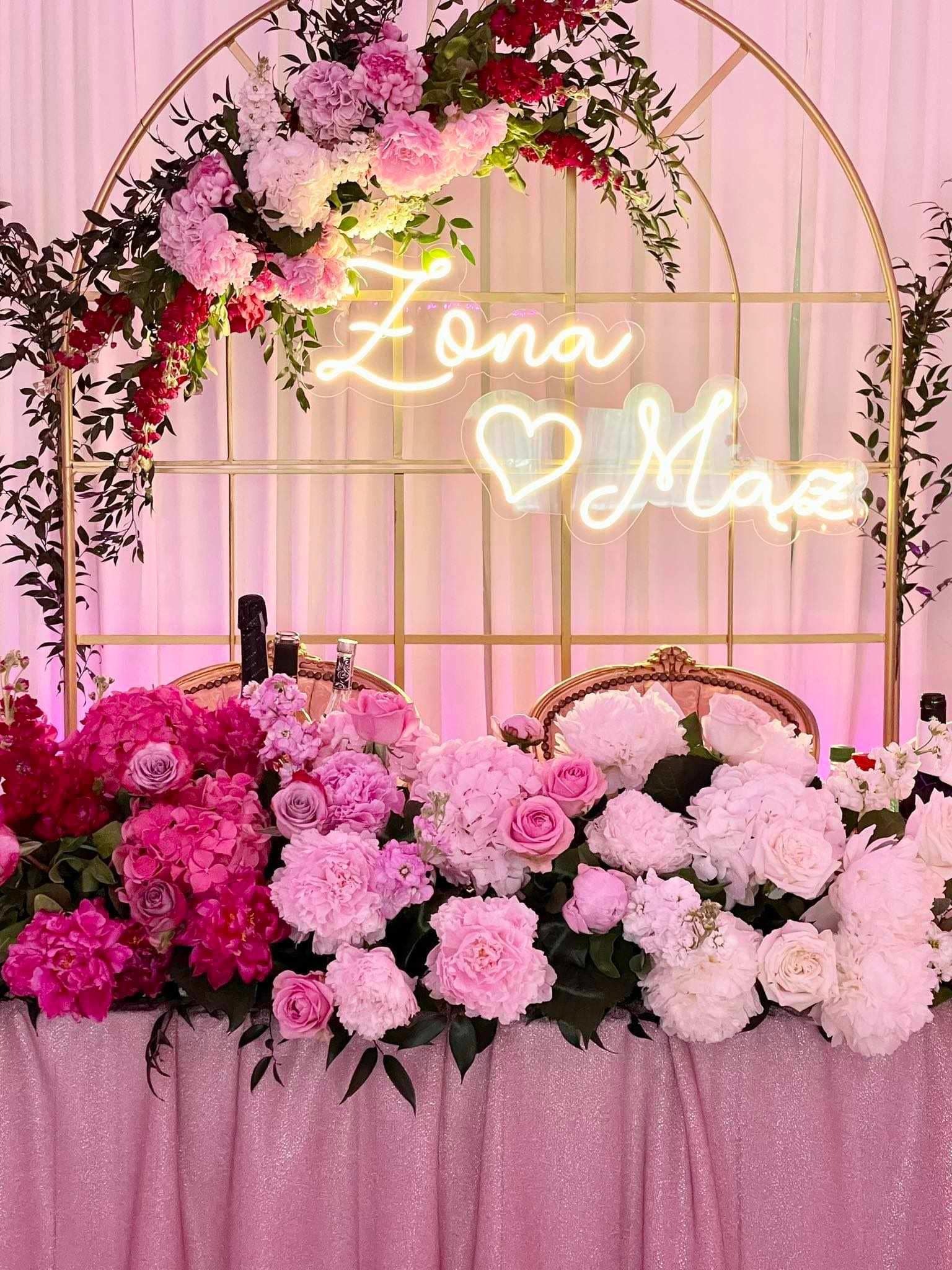 Dekoracja stołu weselnego z kwiatami w odcieniach różu i bieli, neonowym napisem 'Żona ❤️ Mąż' na złotym łuku, butelki szampana w tle.