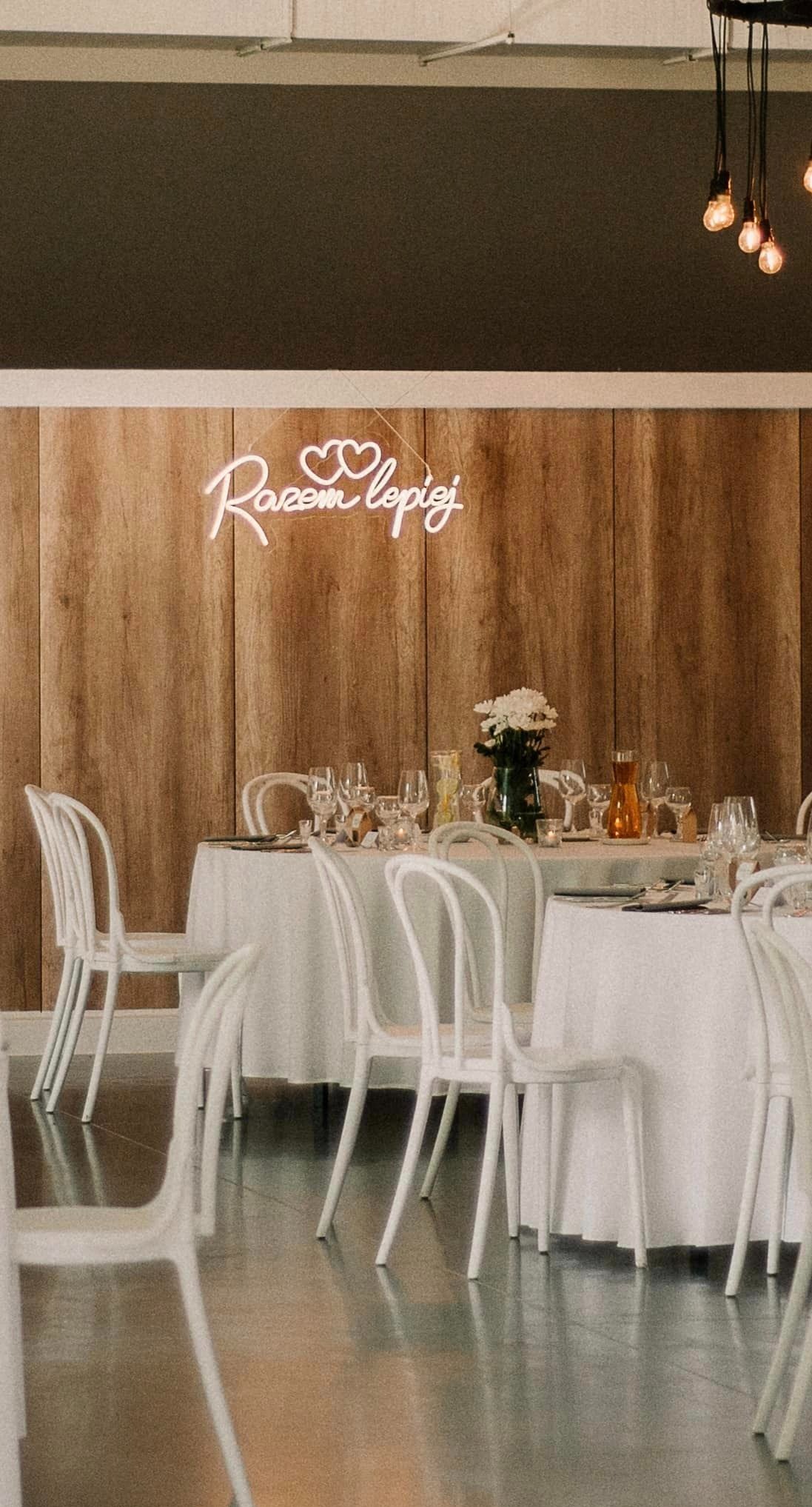 Uroczysty stół weselny z białymi krzesłami, nakryciem, kwiatami w wazonie i dekoracyjnym neonem 'Razem Lepiej' na drewnianym tle.