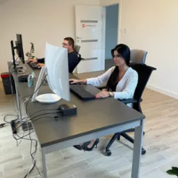 Dwie osoby pracują przy długim, szarym biurku w jasnym biurze. Kobieta w białym kardiganie siedzi przy komputerze, a mężczyzna w okularach pracuje w tle. Widoczne kable, listwa zasilająca i logo...