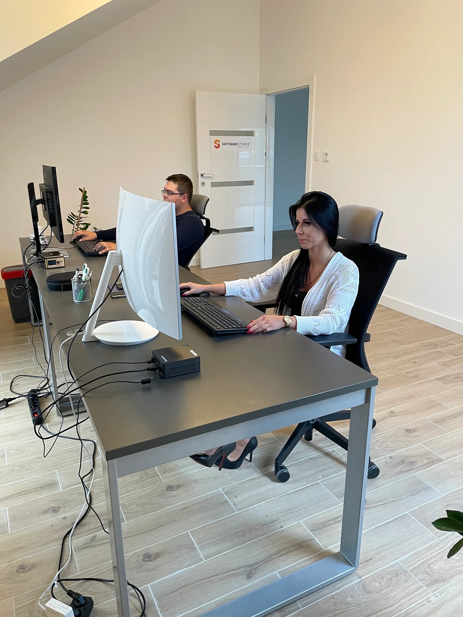 Dwie osoby pracują przy długim, szarym biurku w jasnym biurze. Kobieta w białym kardiganie siedzi przy komputerze, a mężczyzna w okularach pracuje w tle. Widoczne kable, listwa zasilająca i logo...