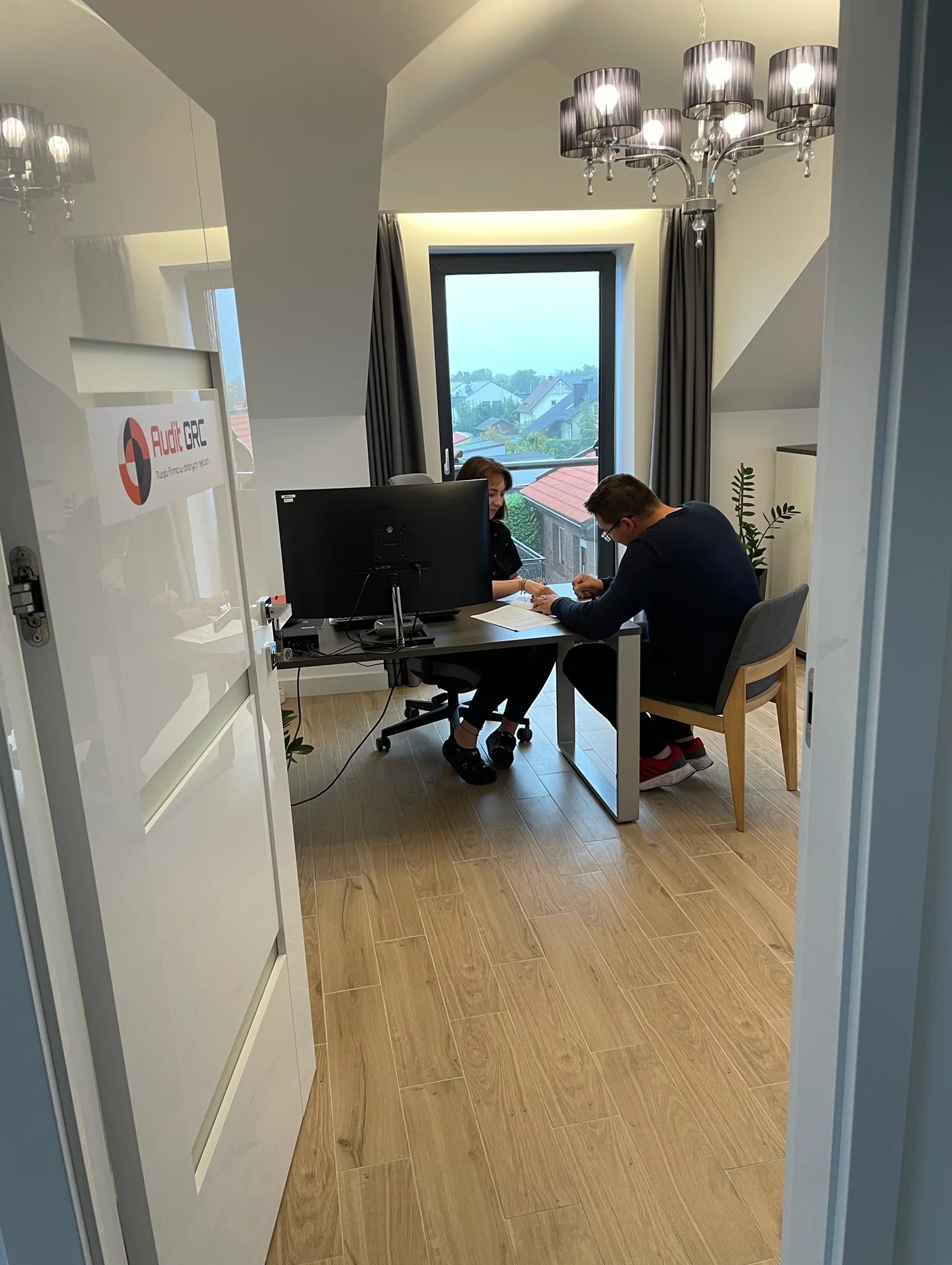 Wnętrze biura z widokiem na dwie osoby pracujące przy biurku, monitor, dokumenty i lampę wiszącą w tle.