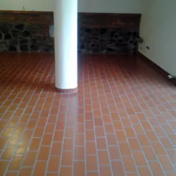 Ułożona podłoga z terakoty w odcieniach brązu, z białą kolumną pośrodku pomieszczenia i fragmentem ściany wyłożonej cegłą i kamieniem w tle.