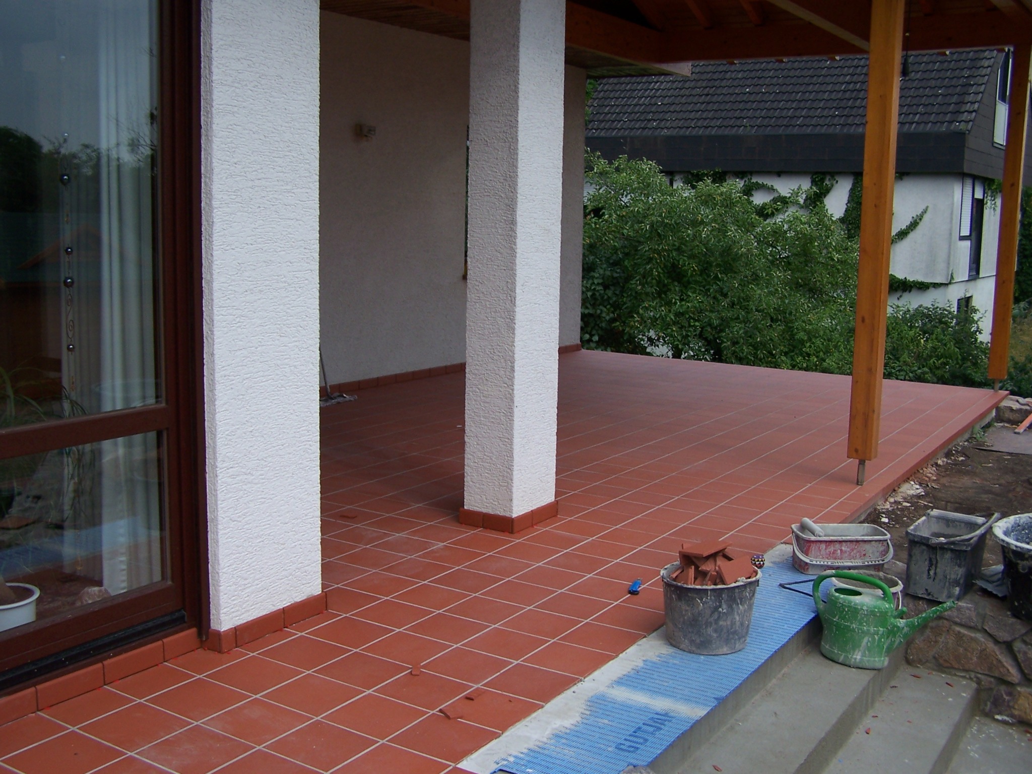 Terracotta płytki podłogowe ułożone na tarasie, widoczne narzędzia glazurnicze i resztki płytek w wiadrach, w tle słupy podtrzymujące zadaszenie i budynek z zielenią.