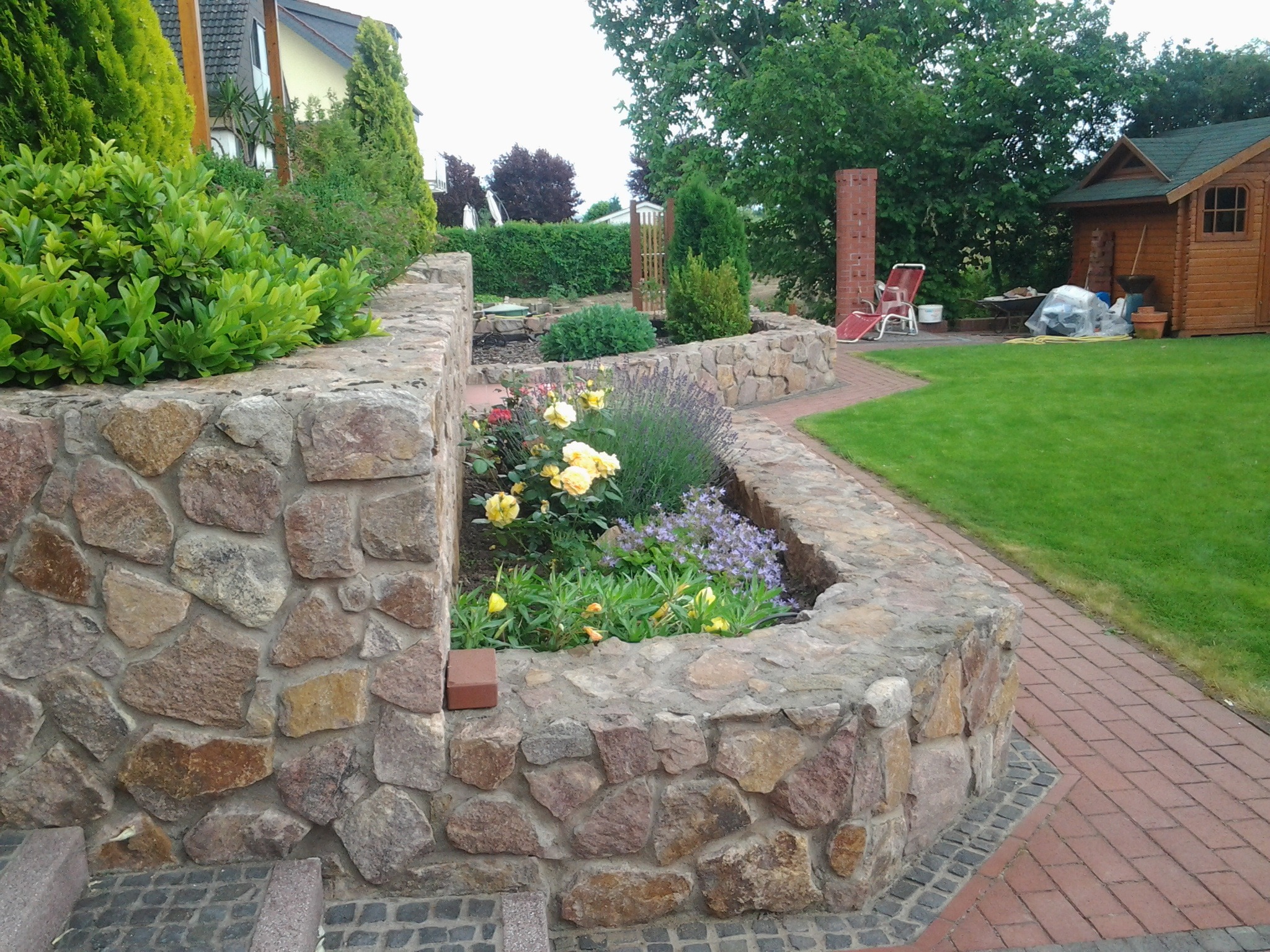 Ogród z kamienną rabatą, w której rosną żółte róże, lawenda i inne kwiaty, z widoczną ścieżką z czerwonej kostki i trawnikiem w tle.