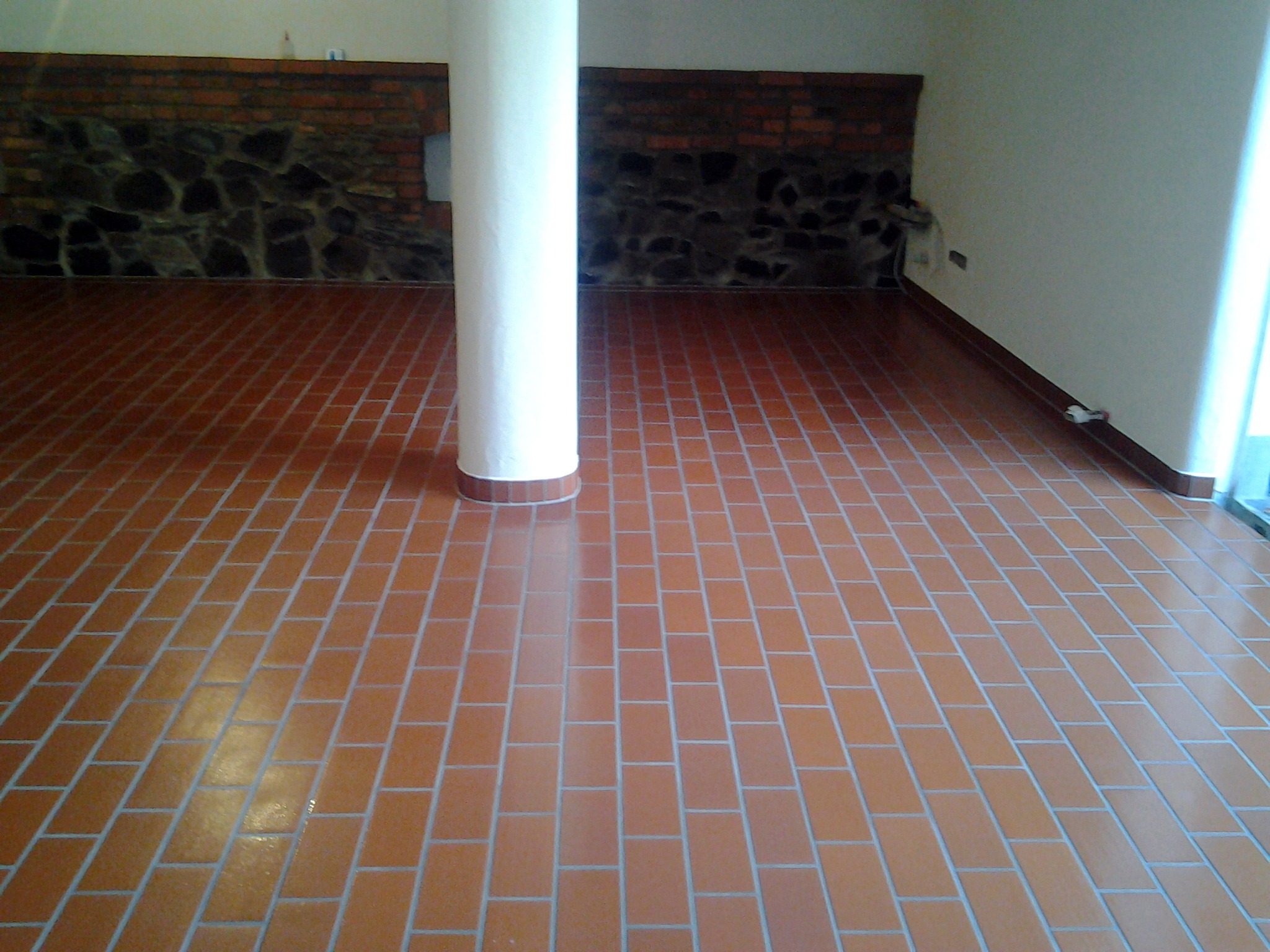Ułożona podłoga z terakoty w odcieniach brązu, z białą kolumną pośrodku pomieszczenia i fragmentem ściany wyłożonej cegłą i kamieniem w tle.