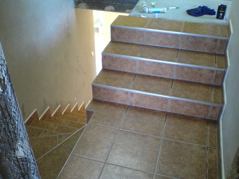 Schody wyłożone brązowymi płytkami ceramicznymi z metalowymi listwami zabezpieczającymi krawędzie, widok z góry, częściowo widoczna ściana i fragment pnia drzewa.