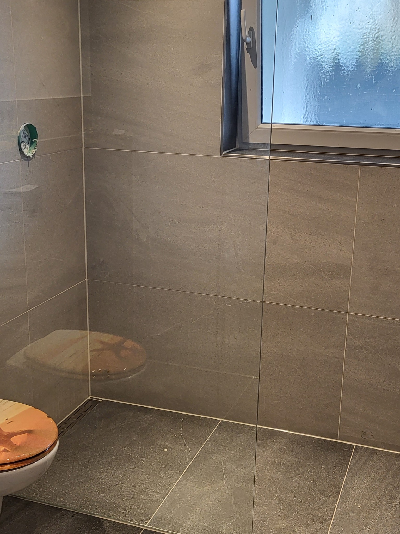 Wykończona łazienka z szarymi płytkami imitującymi kamień, widoczny fragment toalety z deską z motywem morskim oraz okno z widokiem na zewnątrz.