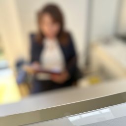 Kancelaria Rzeczoznawcy Majątkowego Katarzyna Włodarczyk-Tomczyk - Kobieta w garniturze przegląda dokumenty za biurkiem w jasnym pomieszczeniu, widok zza blatu.