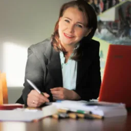 Uśmiechnięta kobieta w biurze, pisząca długopisem na papierze, z laptopem i dokumentami na stole. Portret biznesowy w naturalnym oświetleniu.