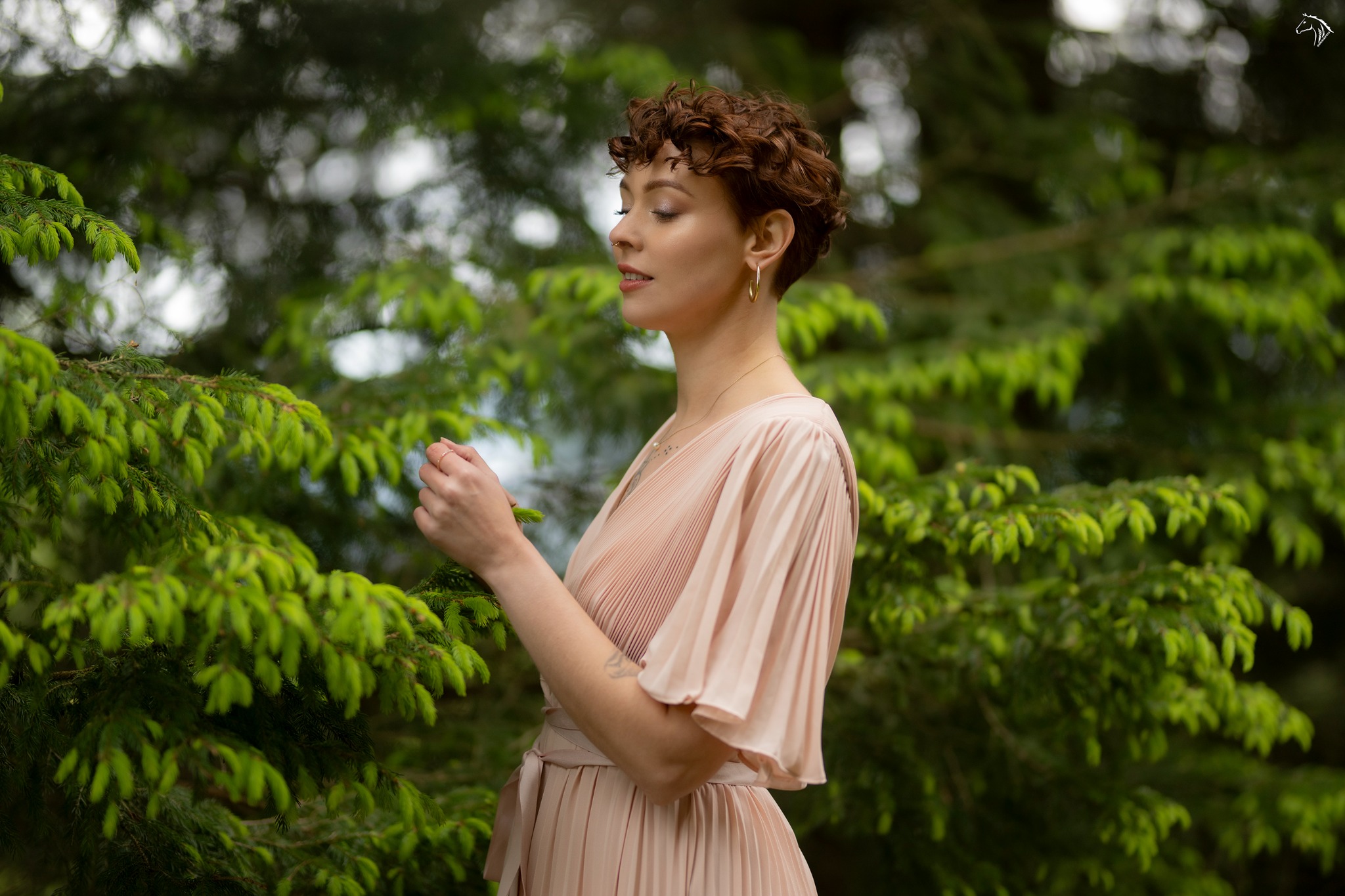Kobieta w pudrowej sukience, z krótkimi kręconymi włosami, stoi profilem na tle zielonych gałązek drzew iglastych, trzymając delikatnie gałązkę w dłoni.