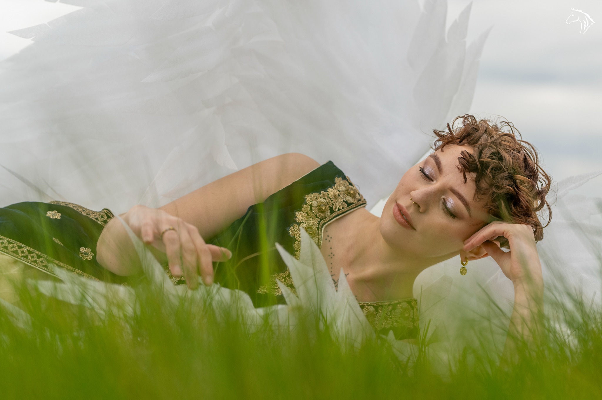 Kobieta w zielonej sukni z haftem leży na trawie, otulona białym, zwiewnym materiałem, z zamkniętymi oczami i subtelnym makijażem.